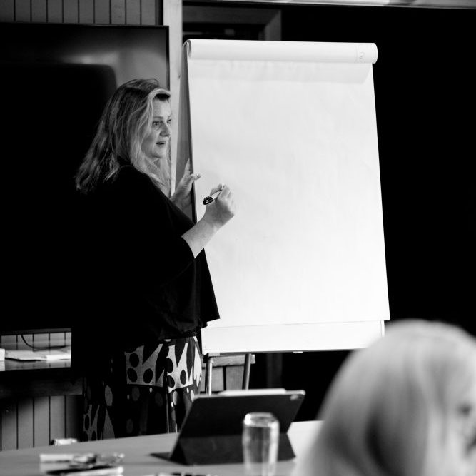 Black and white image of Tess Crawley, psychologist and business coach, teaching a group of professionals. She is standing at a white board ready to take notes.