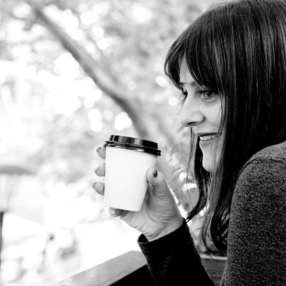 Black and white photo of Tess Crawley, psychologist and business coach, looking out over the Yarra River, holding a takeaway cup of coffee and smiling.